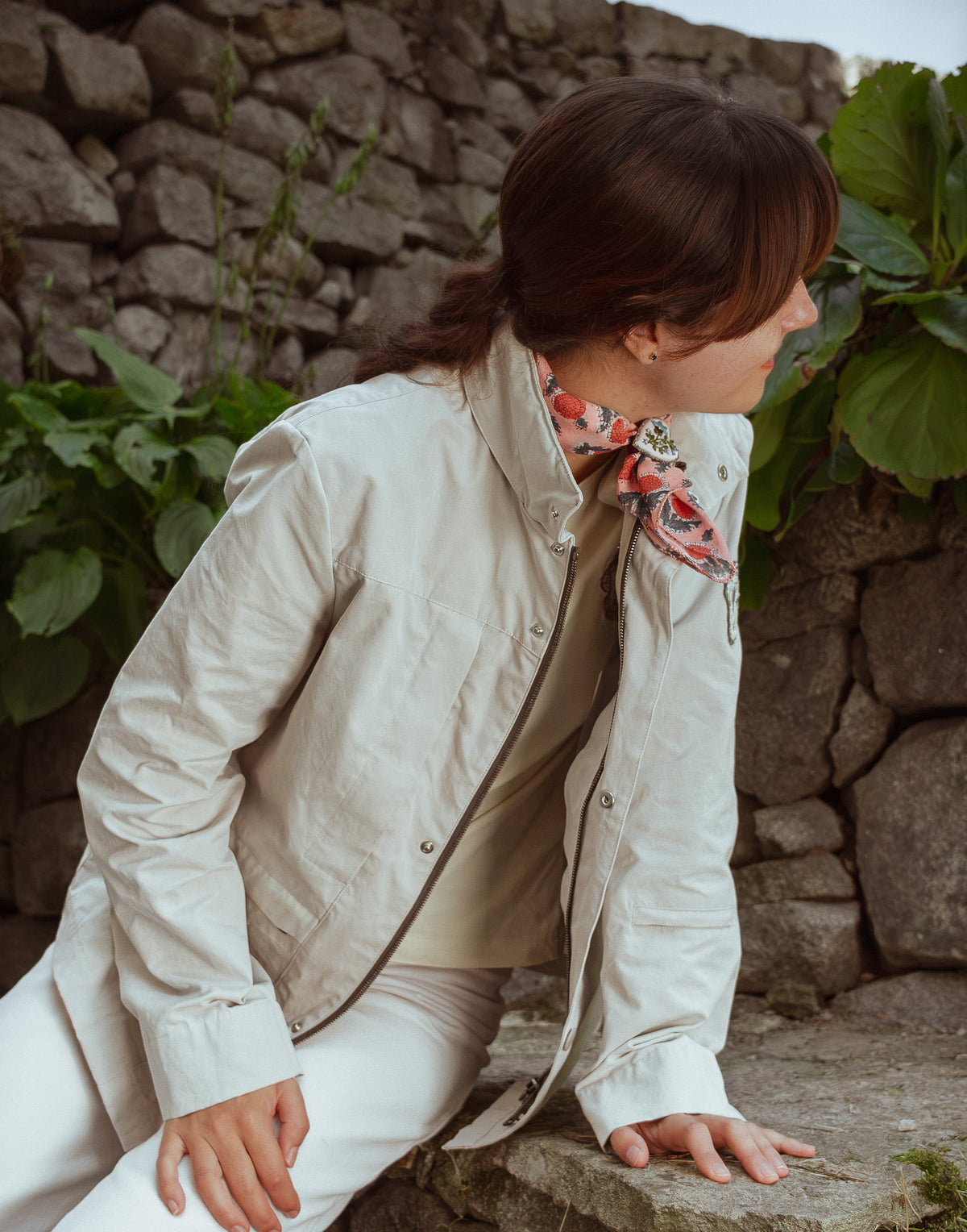 A woman wearing a light beige jacket, beige top, and white pants, leaning on a stone wall outdoors with green plants in the background.