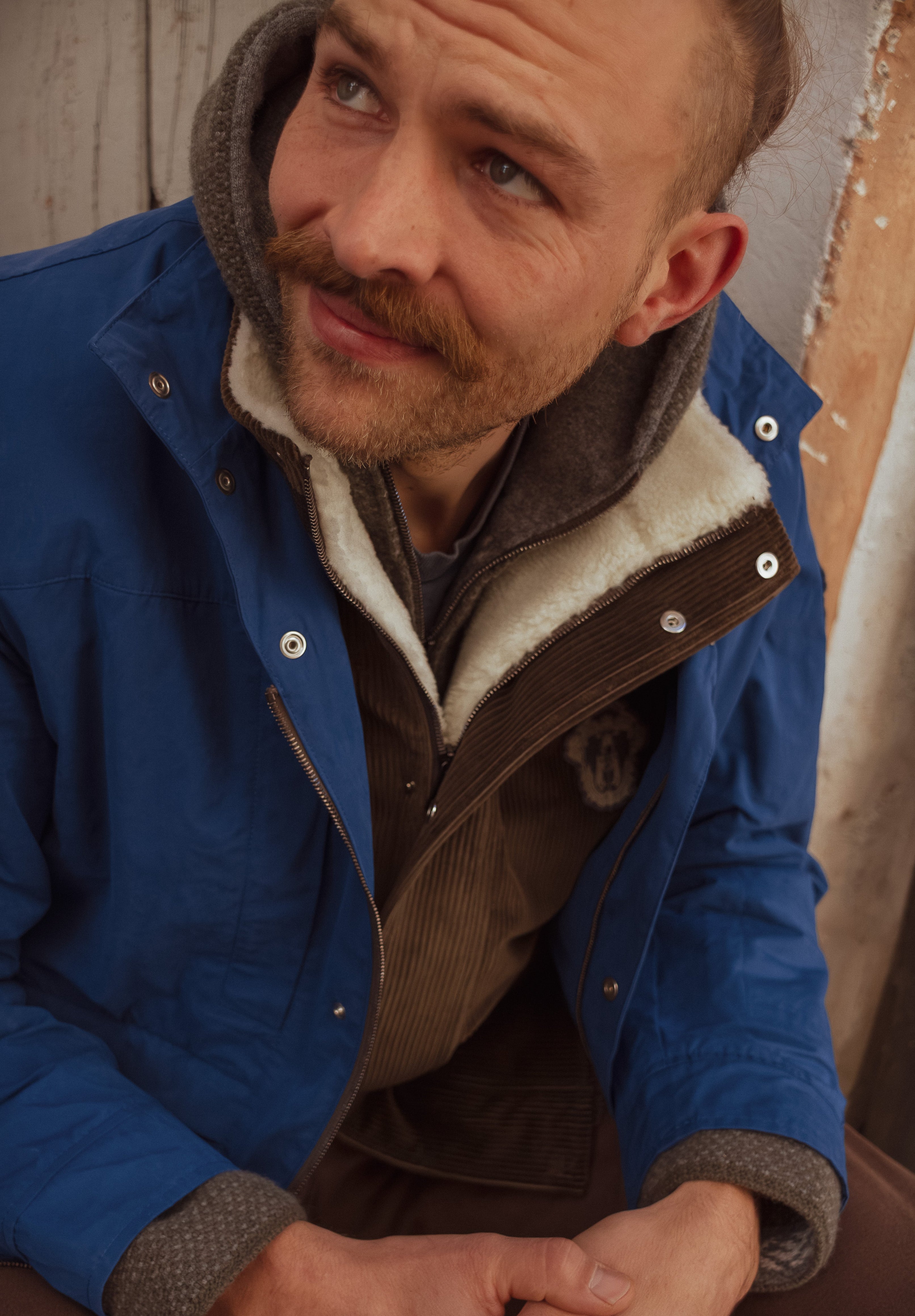 A close-up lifestyle shot of a man wearing a blue hooded jacket with a cream sherpa lining, layered over a brown sweater.