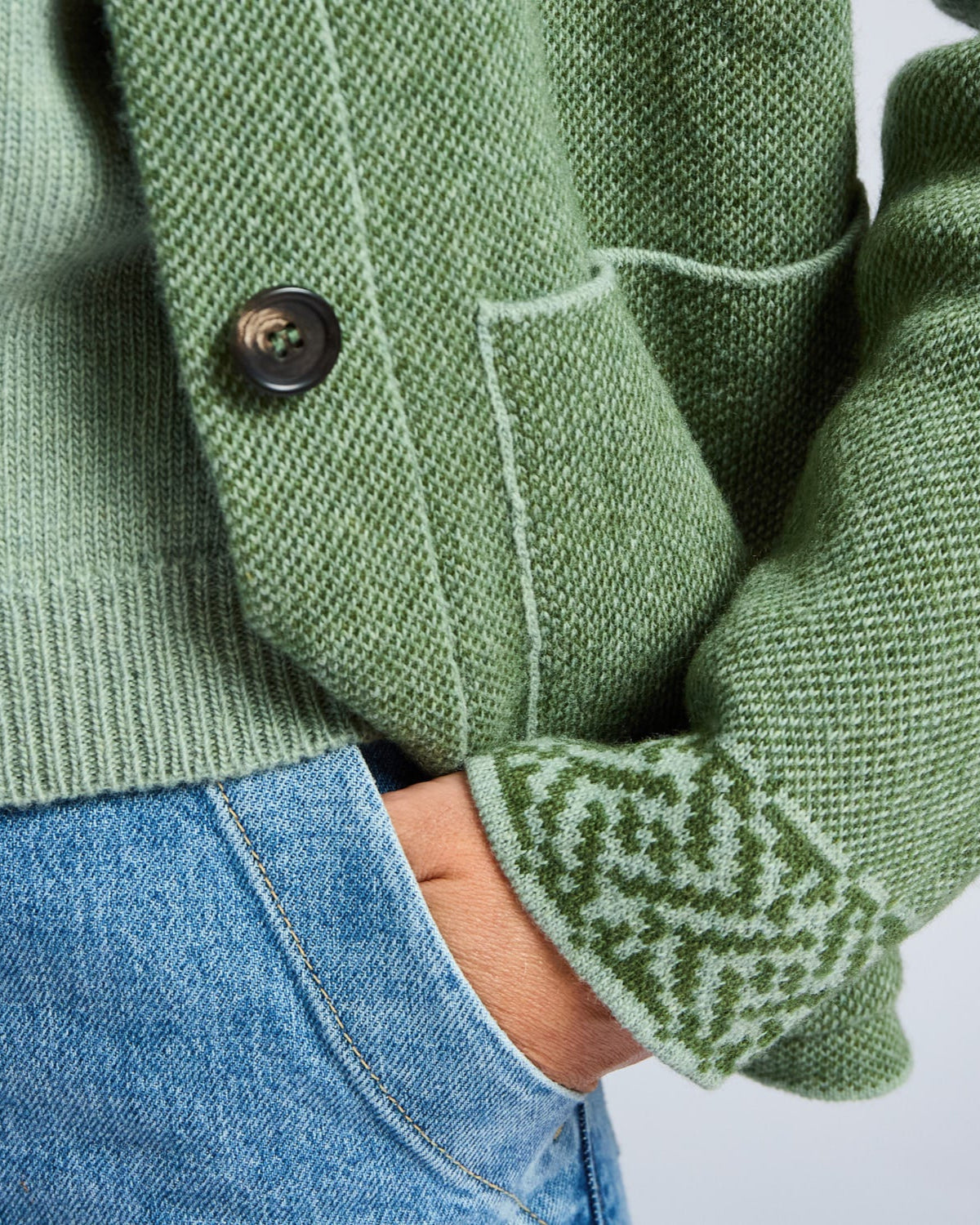 Green knitted cardigan with patterned cuffs and a button, worn over blue jeans.