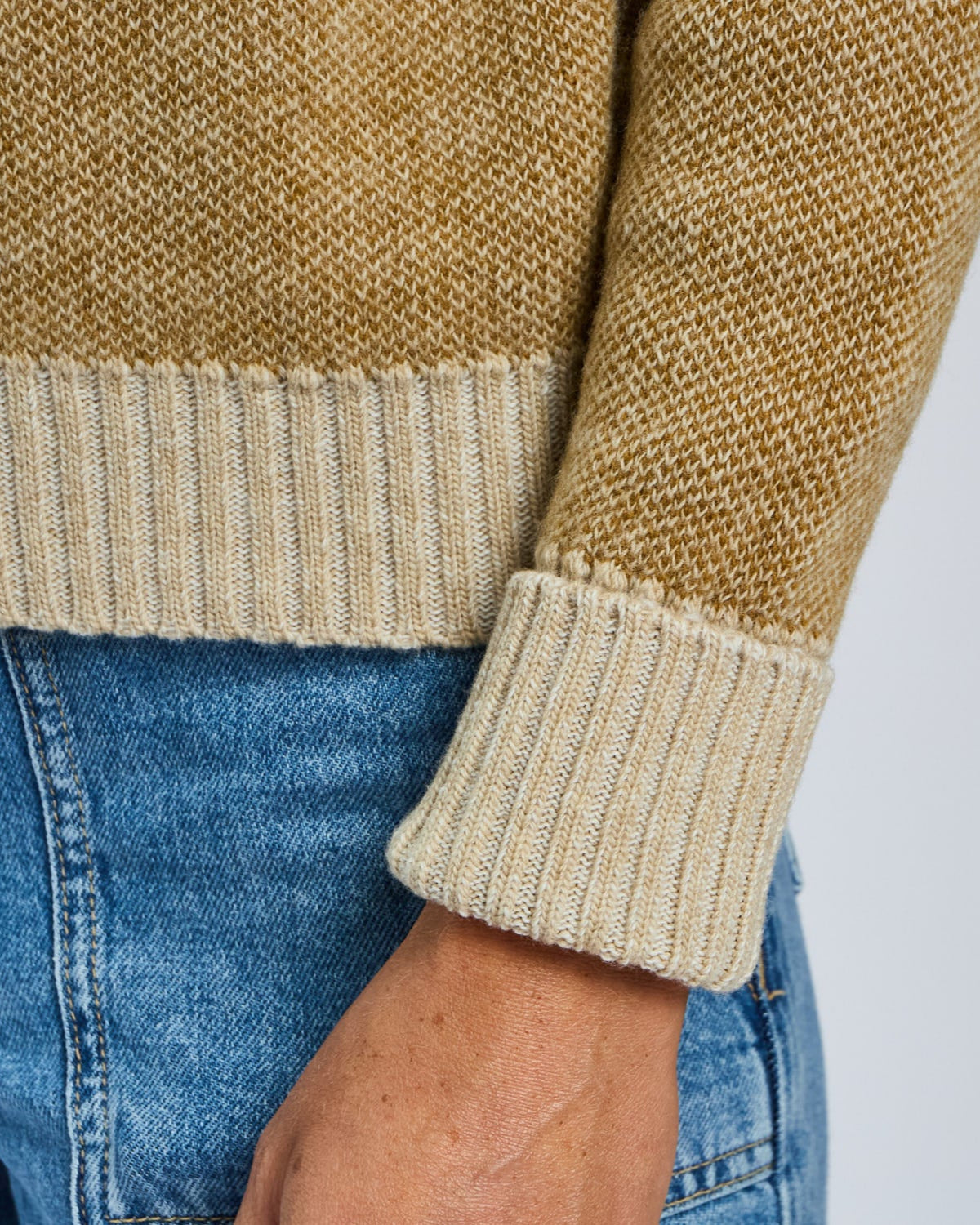 Close-up of a person wearing a beige sweater and blue jeans on a light gray background