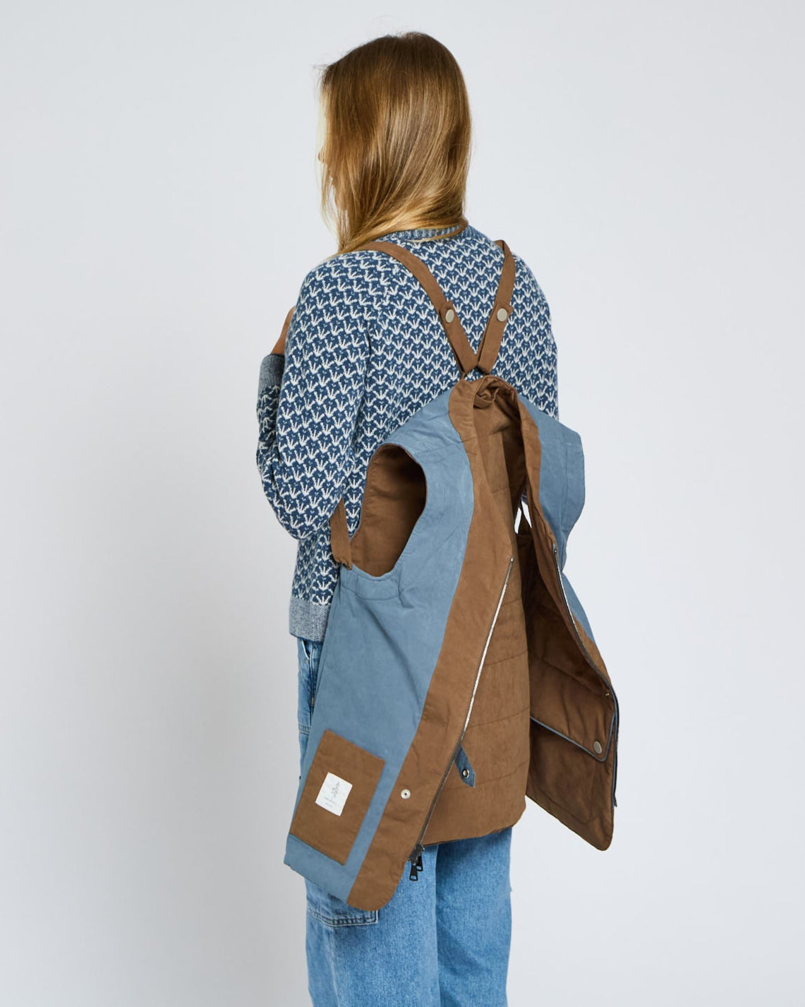 Person wearing a blue patterned shirt and brown apron overalls on a white background