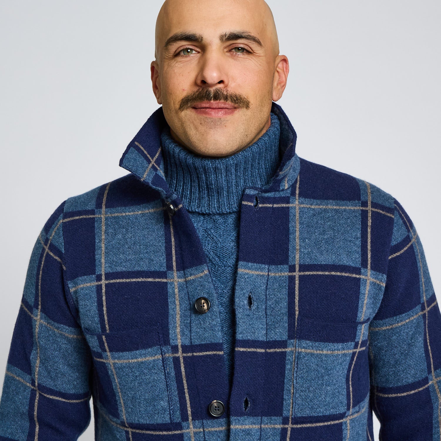 Man wearing a blue checkered coat over a turtleneck sweater against a plain background
