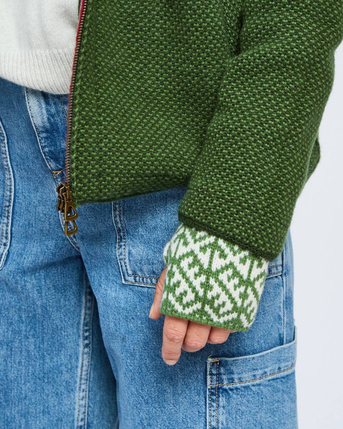 Green knitted sleeve and patterned green and white glove held by a person wearing blue jeans.
