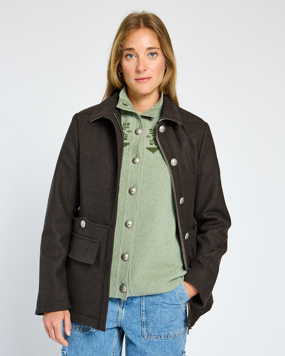 Woman wearing a dark brown coat over a green shirt and blue jeans on a white background