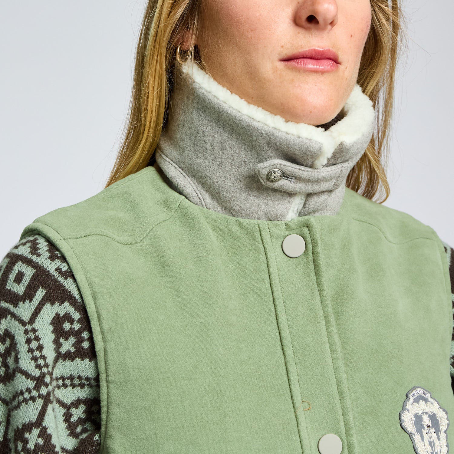 Person wearing a green vest with patterned sleeves and a gray scarf against a white background