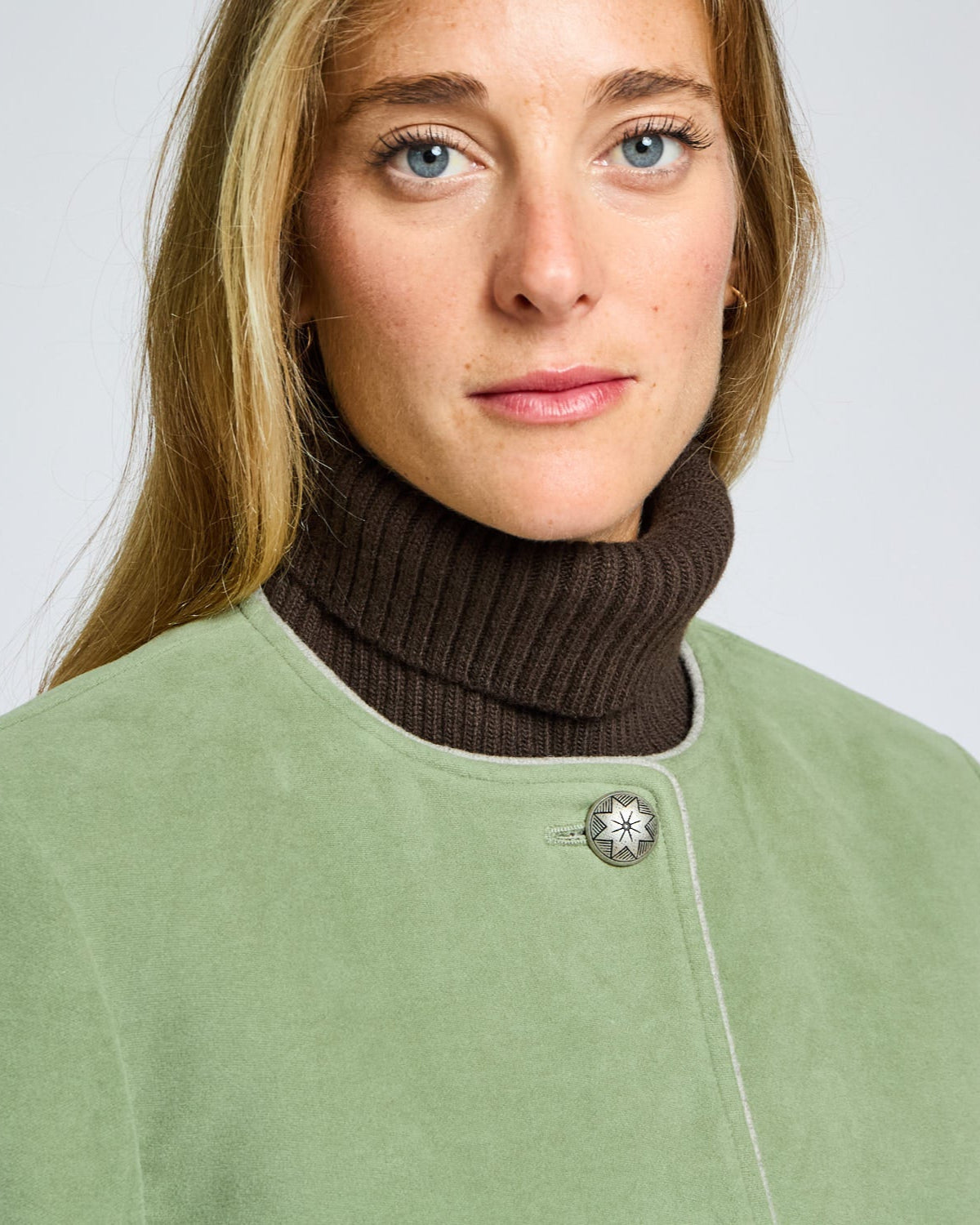 Person wearing a green coat with a brown turtleneck underneath on a plain background