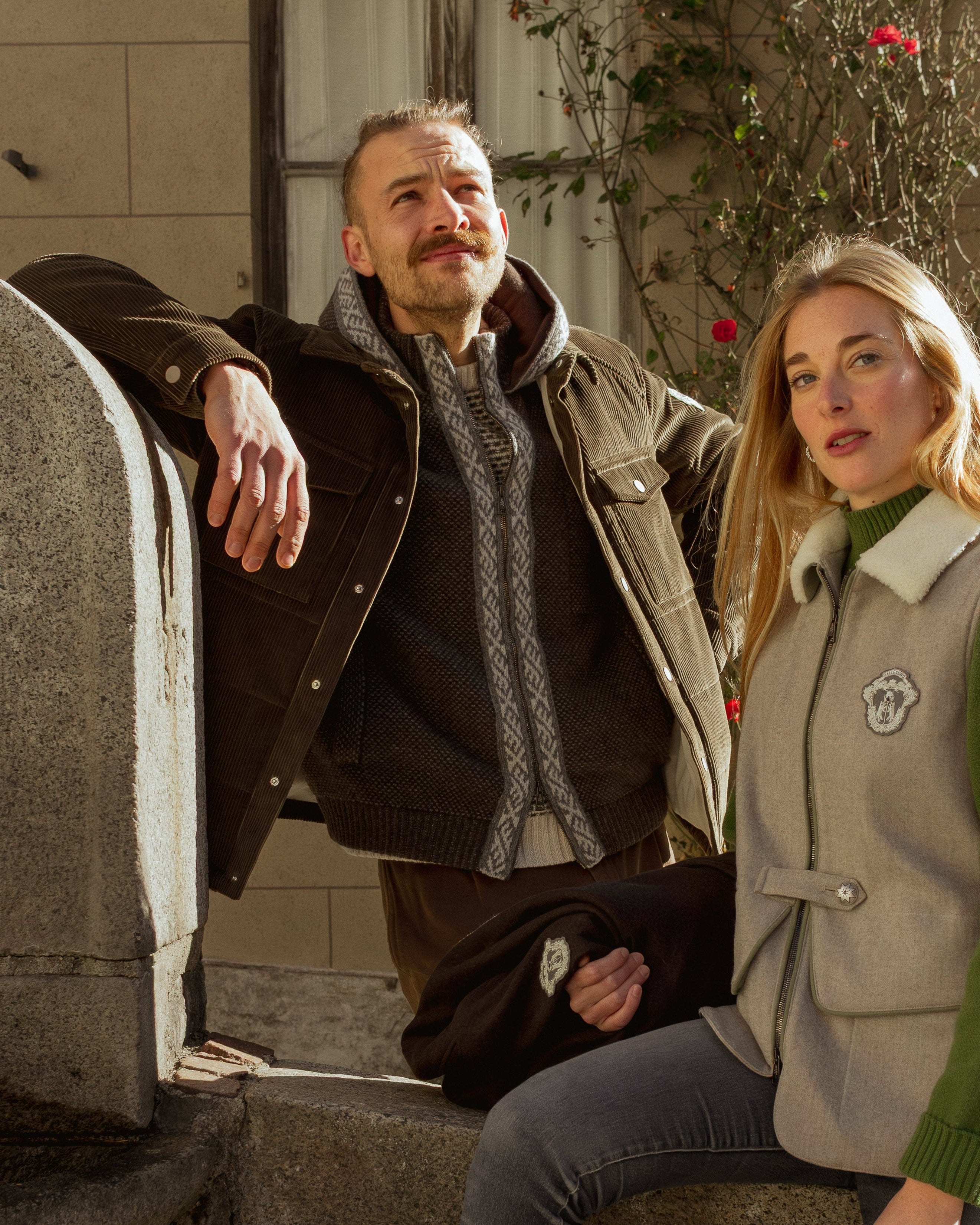 Two people leaning against a stone wall with a building and tree in the background