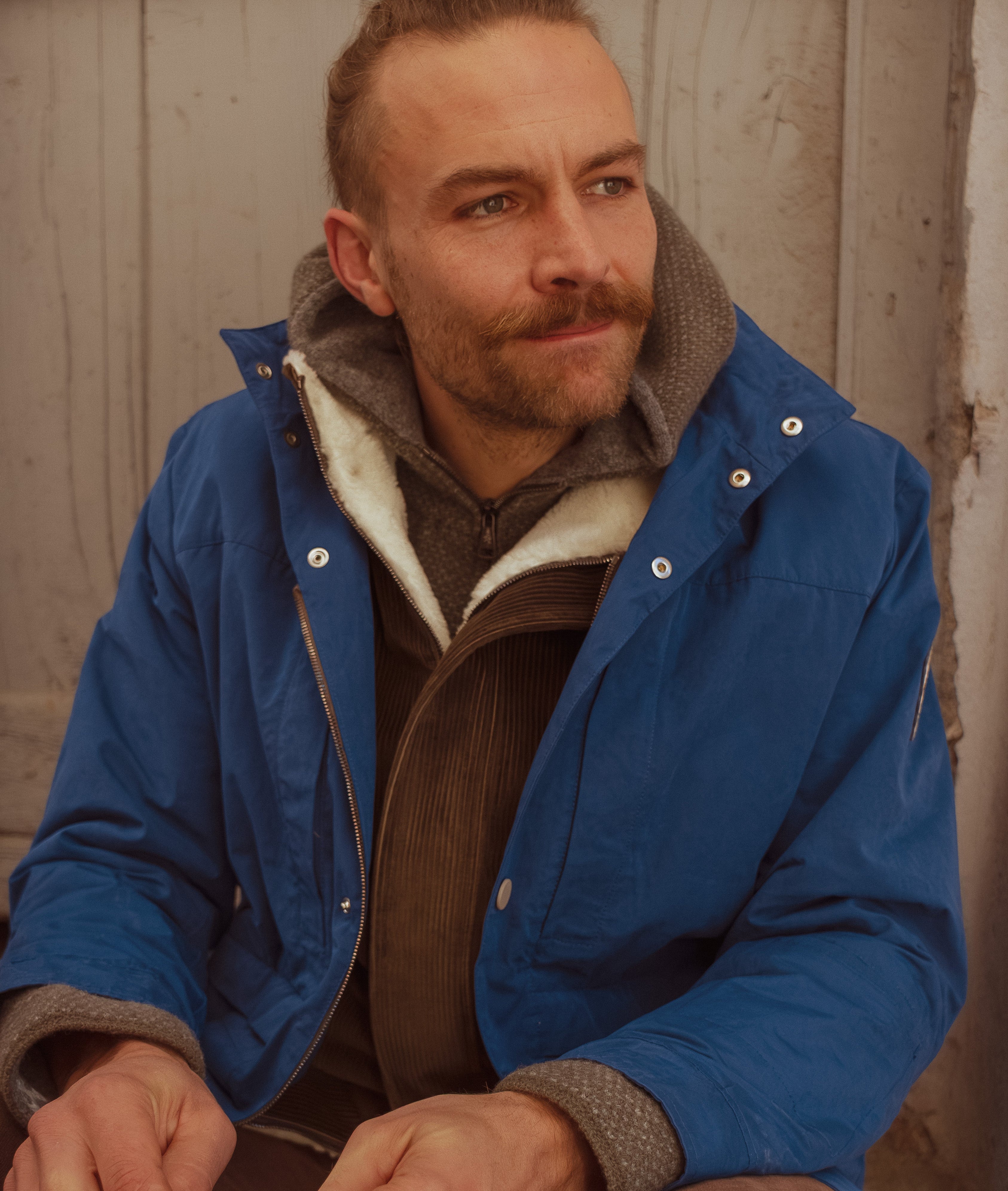 A man wearing a blue hooded jacket with a brown corduroy shirt and brown pants, sitting against a rustic wooden wall.