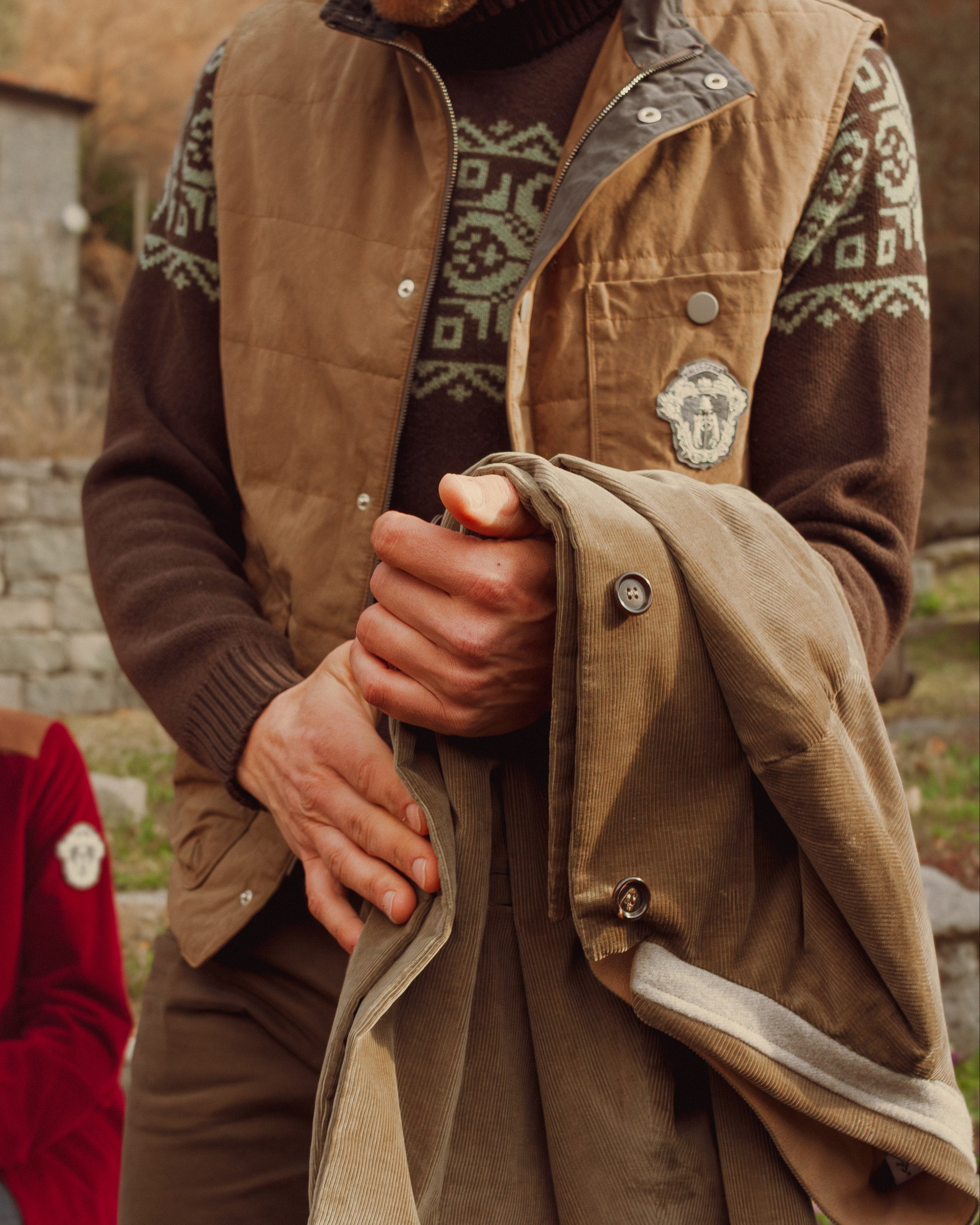 Person wearing a brown jacket with patterned sleeves holding a beige coat outdoors.