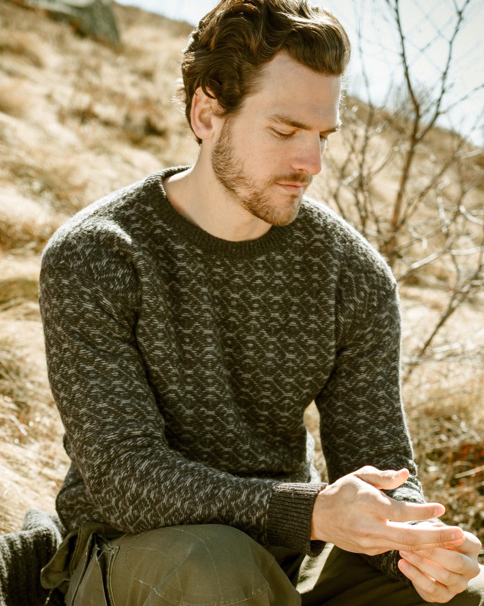 A man wearing the Cima Bianca NIK - Wool Crewneck Sweater with Intarsia sits outside on dry grass beside a blanket, dressed in green pants and brown boots. Sunlight casts a warm glow as he looks down at his hands.