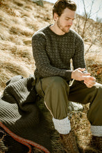 Load image into Gallery viewer, A man wearing the Cima Bianca NIK - Wool Crewneck Sweater with Intarsia sits outside on dry grass beside a blanket, dressed in green pants and brown boots. Sunlight casts a warm glow as he looks down at his hands.
