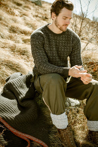 A man wearing the Cima Bianca NIK - Wool Crewneck Sweater with Intarsia sits outside on dry grass beside a blanket, dressed in green pants and brown boots. Sunlight casts a warm glow as he looks down at his hands.