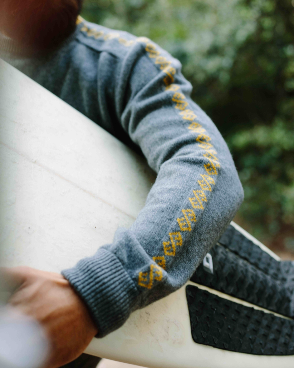 Person wearing a gray sweater with yellow patterns, holding a surfboard outdoors.