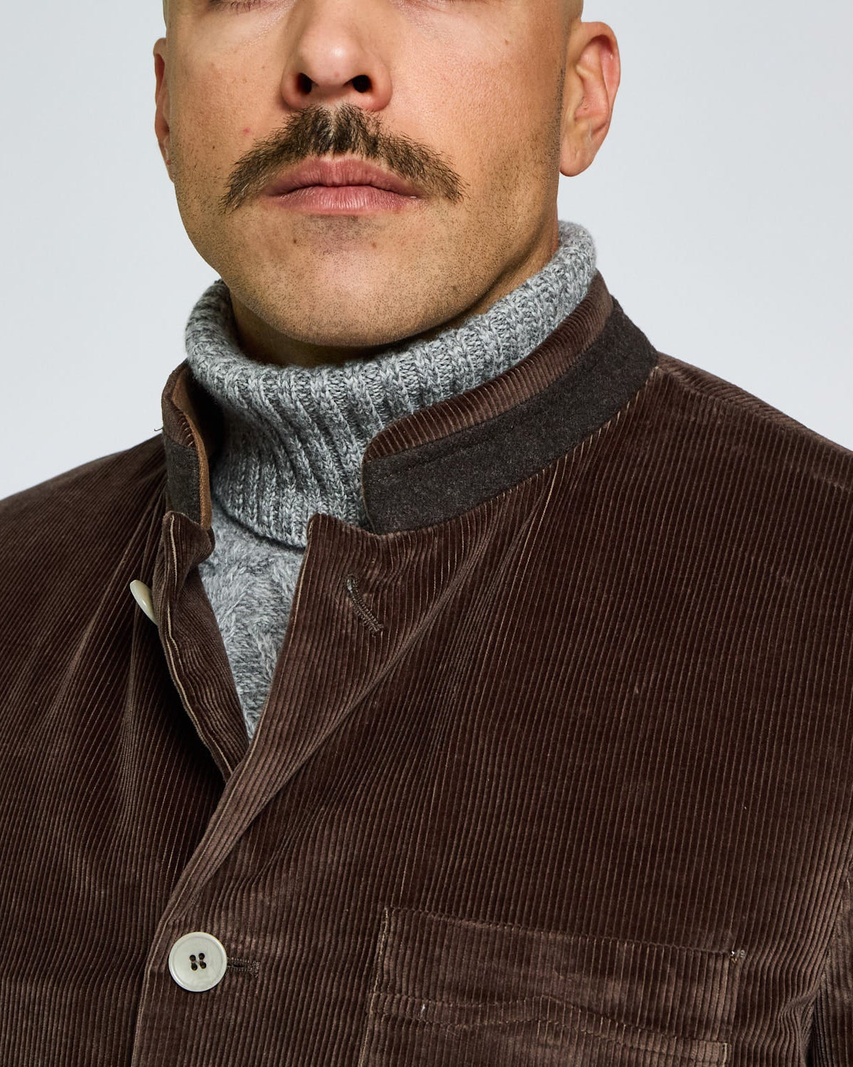 Man wearing a brown corduroy jacket over a gray turtleneck sweater on a light gray background