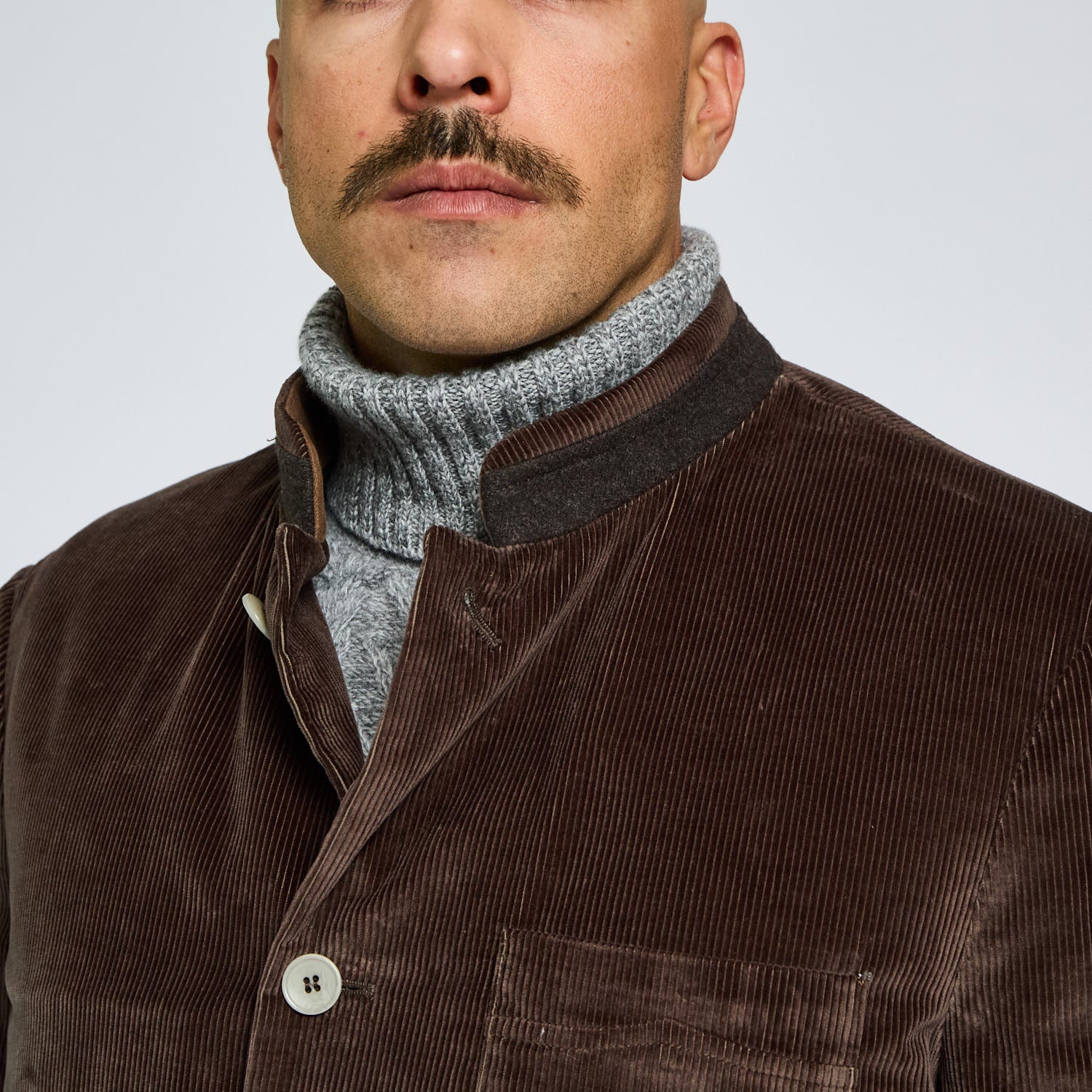 Man wearing a brown corduroy jacket over a gray turtleneck sweater on a light gray background