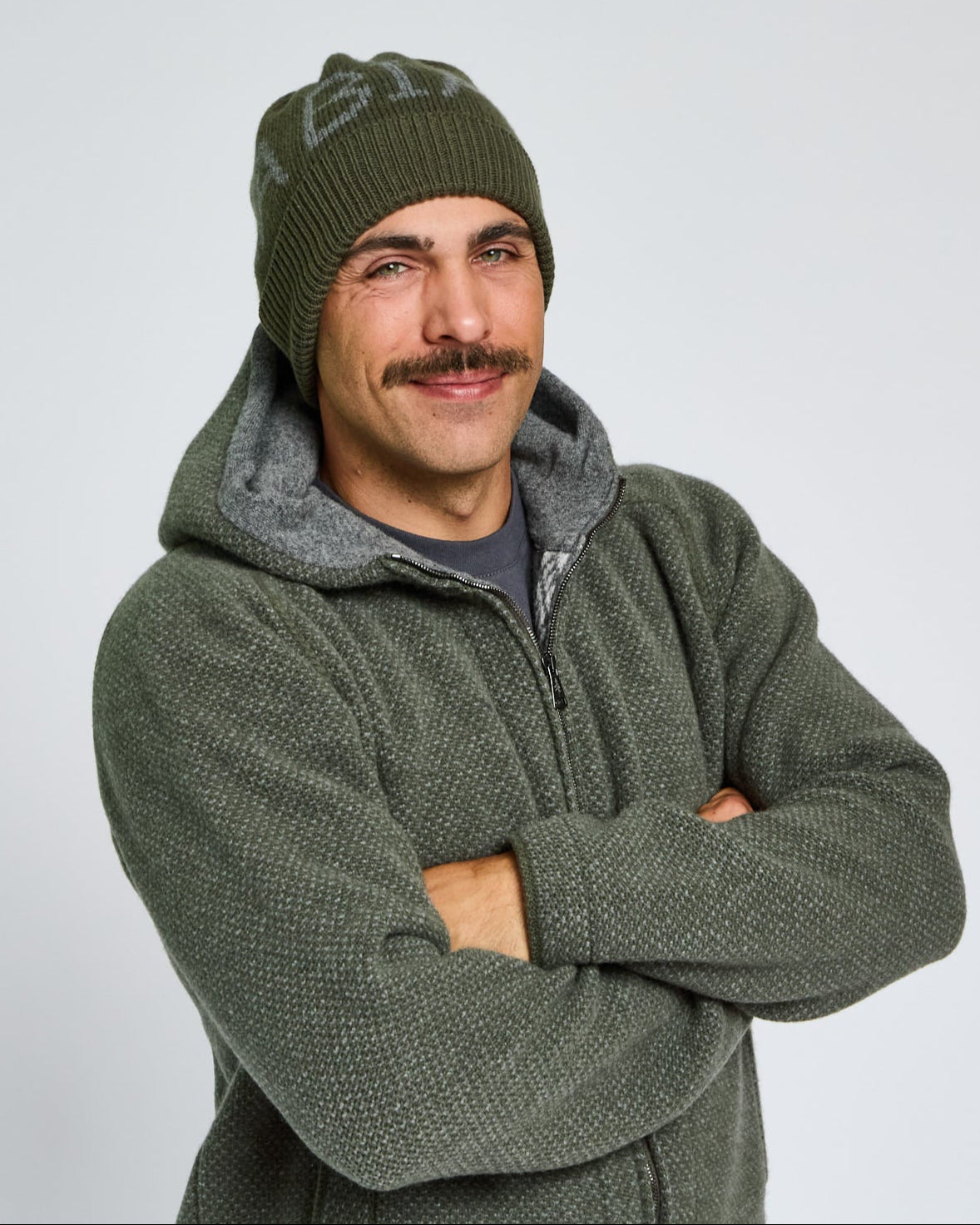 Man wearing a green knit beanie and matching jacket with arms crossed on a light gray background