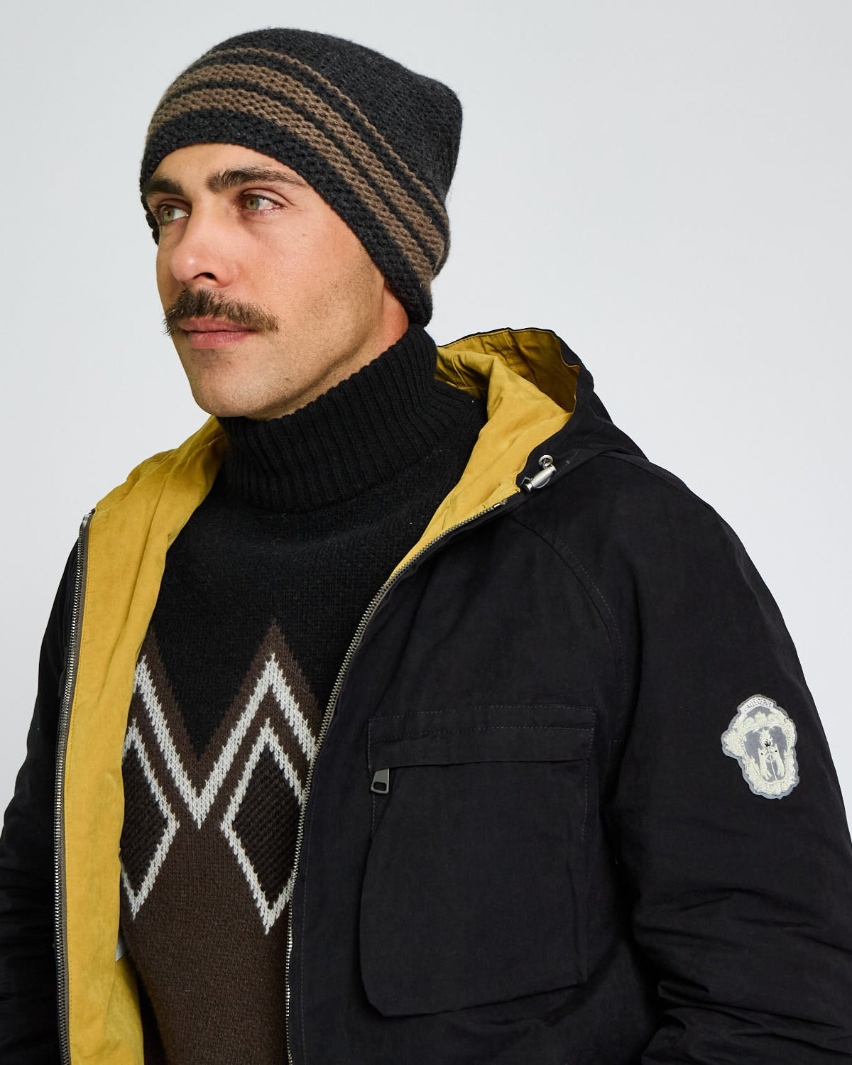 Man wearing a black jacket with yellow lining, black knit beanie, and patterned sweater on a light gray background