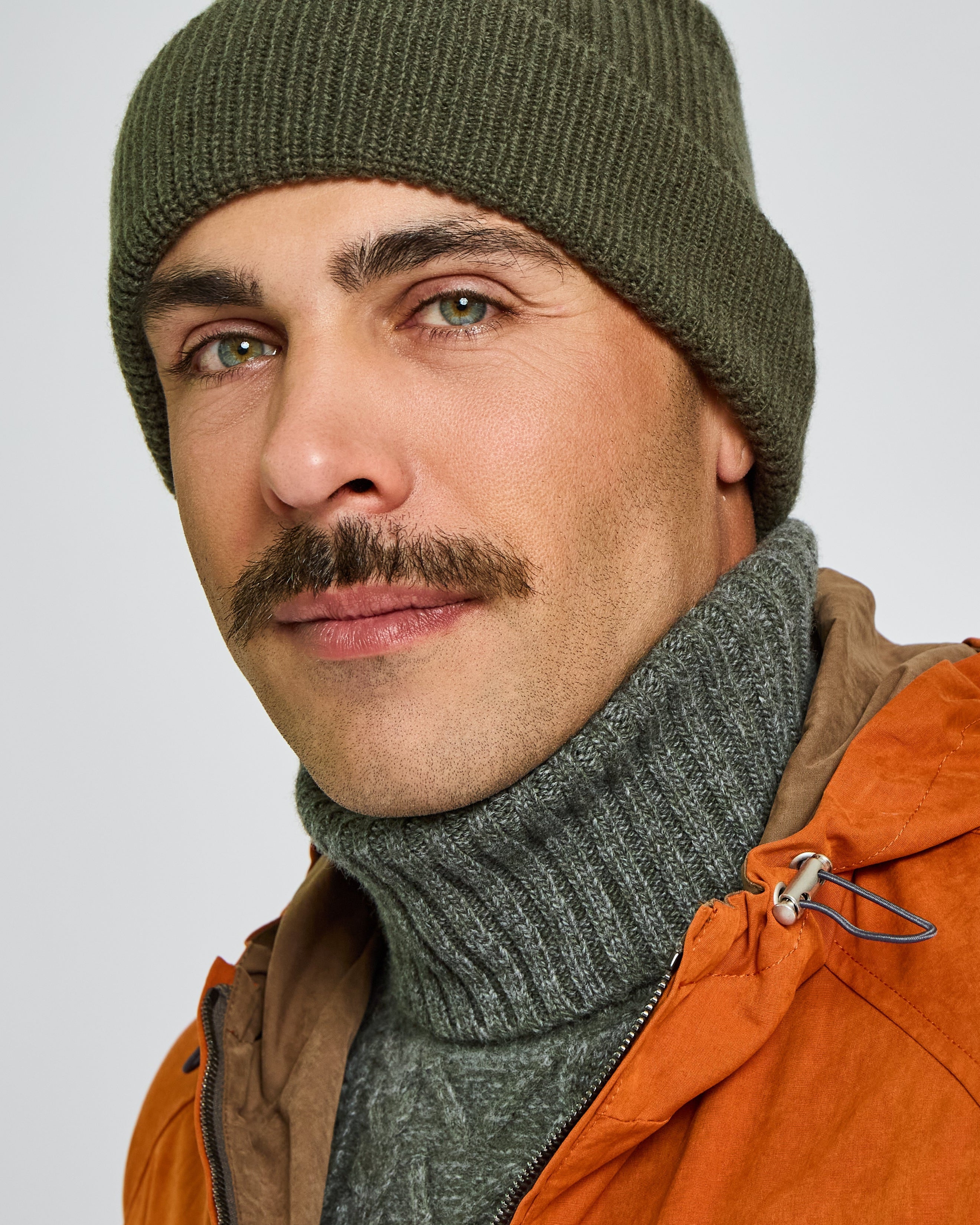 Man wearing an orange jacket, green beanie, and gray turtleneck against a plain background