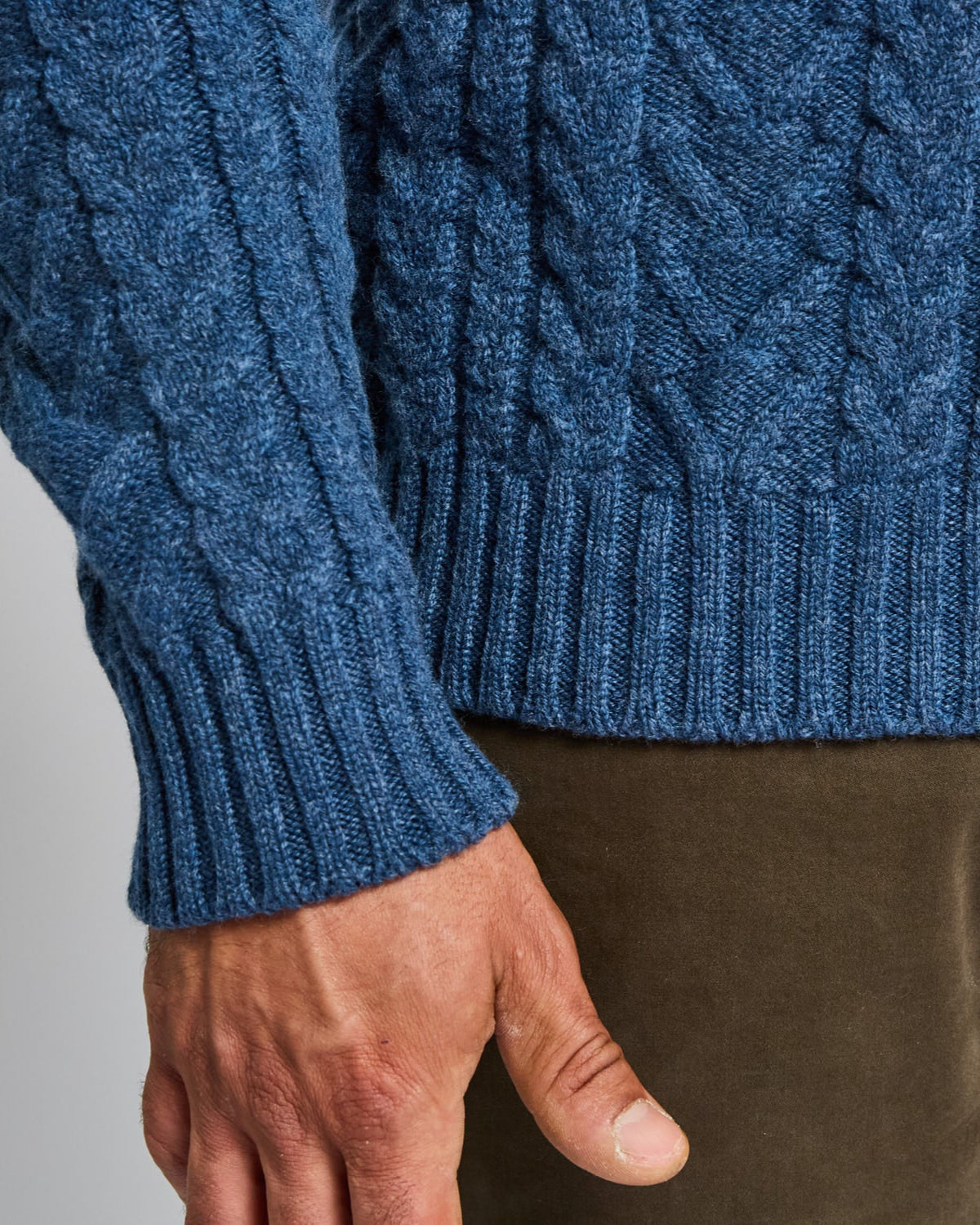 Close-up of a blue cable knit sweater with a hand wearing it against a gray background