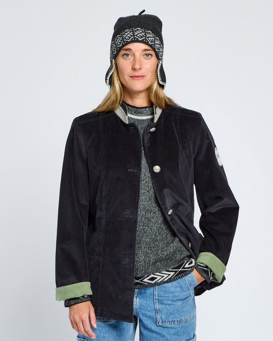 Person wearing a black jacket with green accents, blue jeans, and a patterned beanie on a light gray background