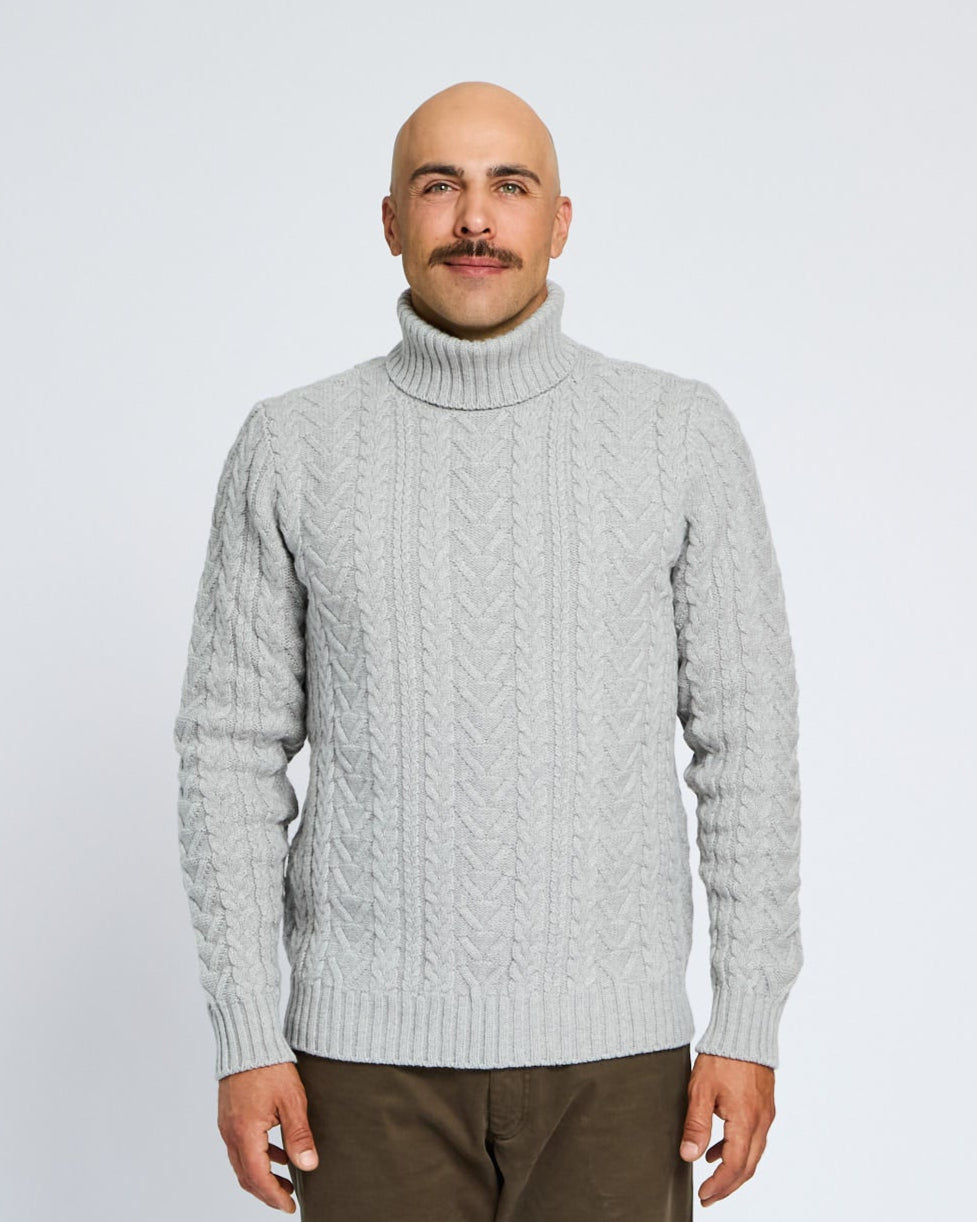 Man wearing a light gray turtleneck sweater and brown pants on a white background