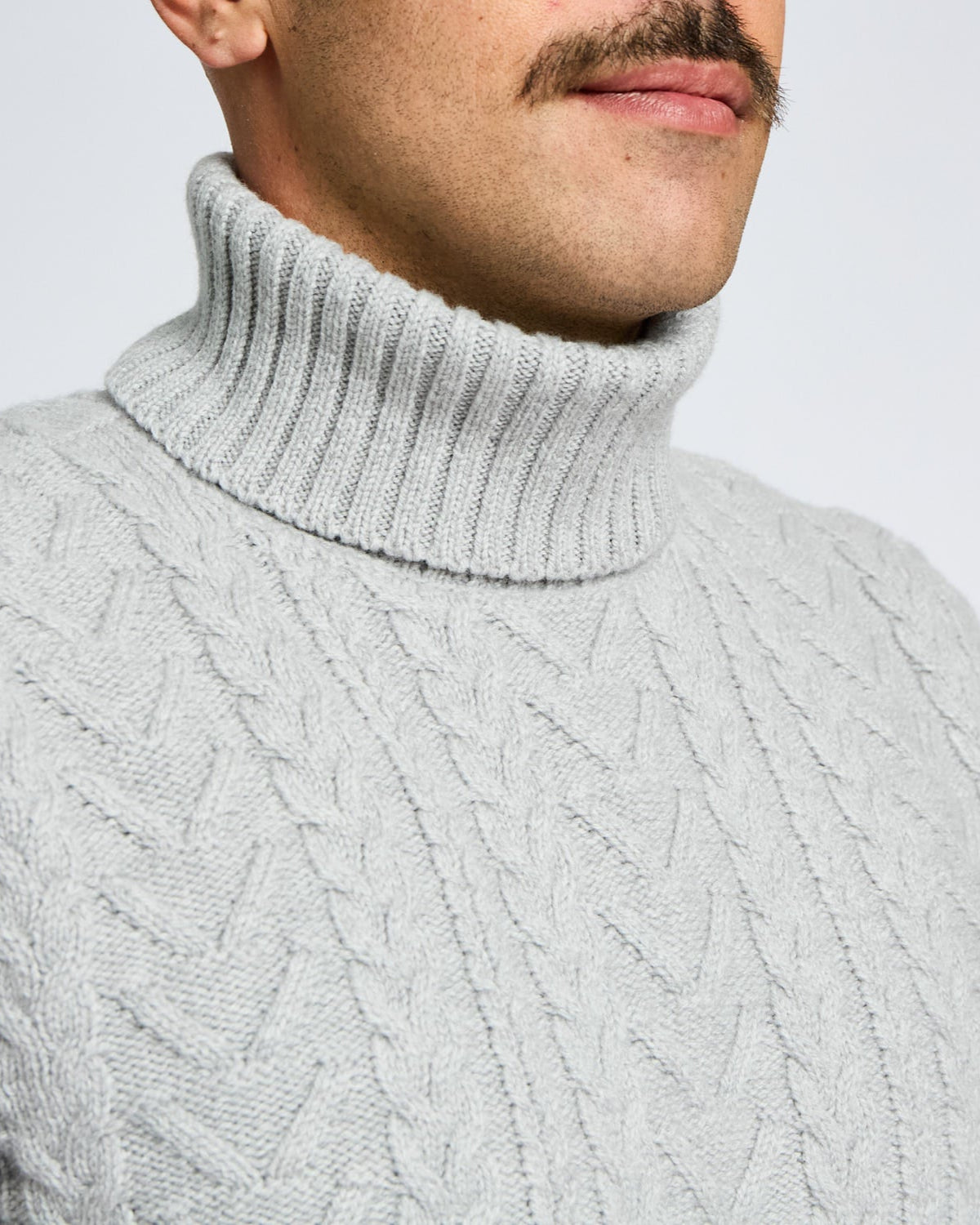 Close-up of a person wearing a gray cable knit turtleneck sweater against a light gray background