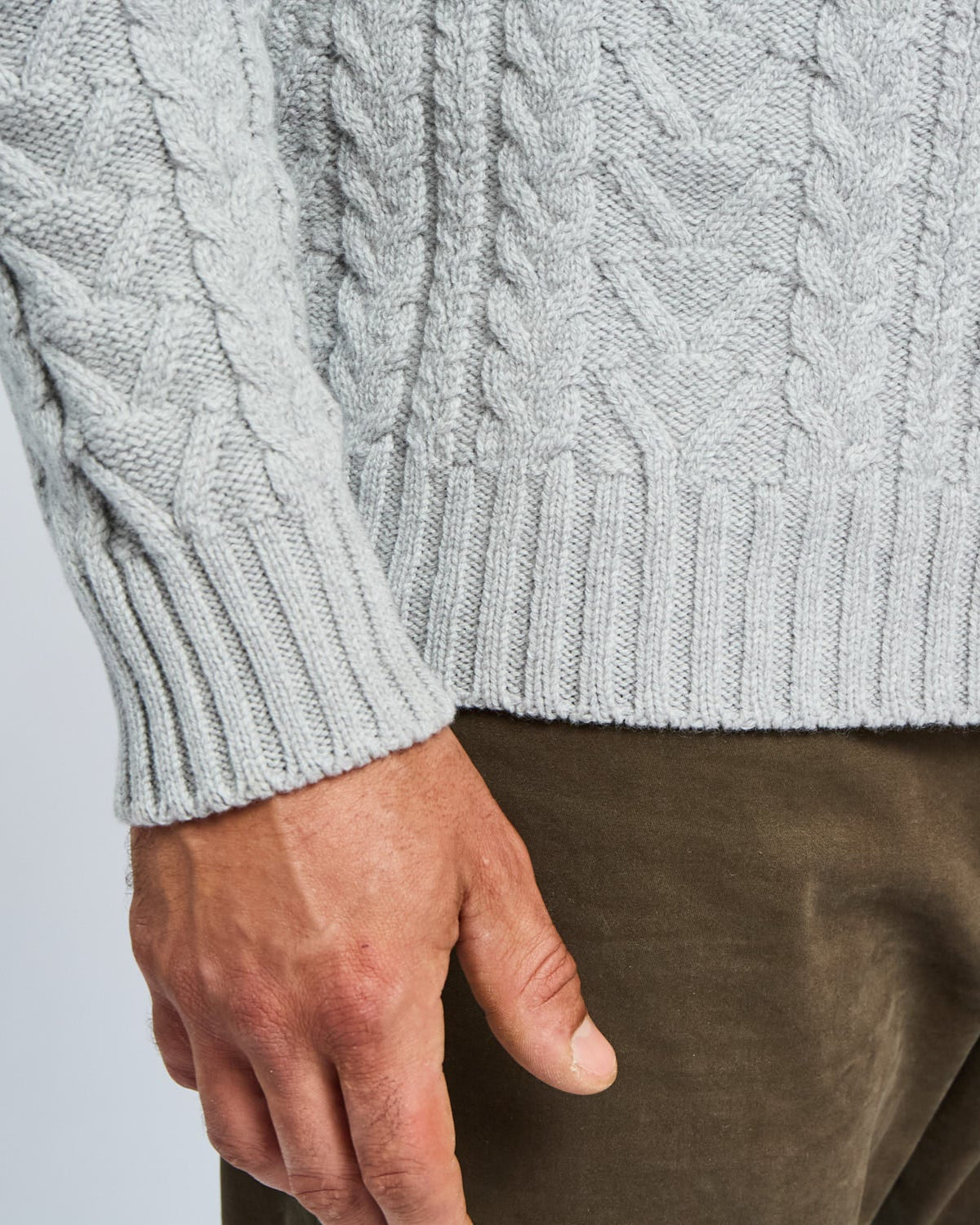 Close-up of a person wearing a gray cable knit sweater and brown pants on a light gray background