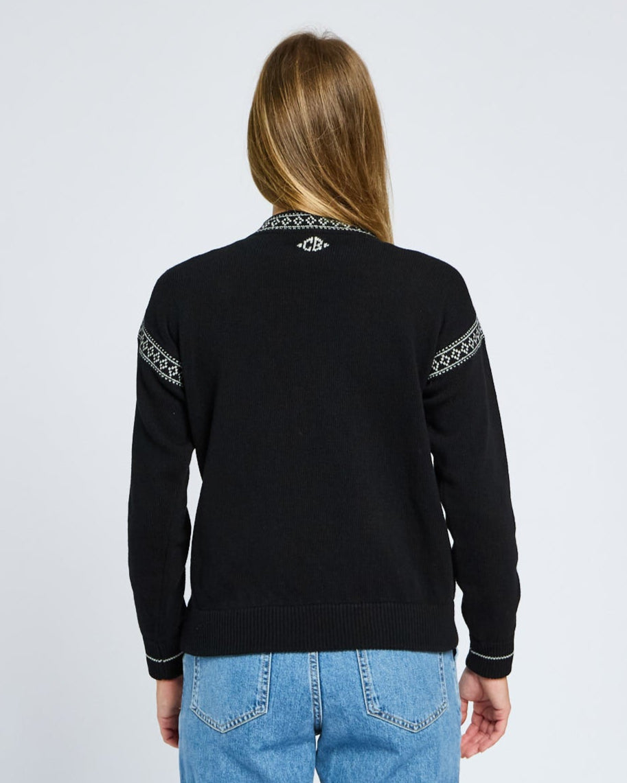 Person wearing a black sweater with white patterns on sleeves and collar, standing against a light gray background.