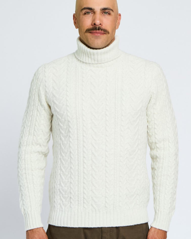 Man wearing a cream sweater and brown pants against a light gray background