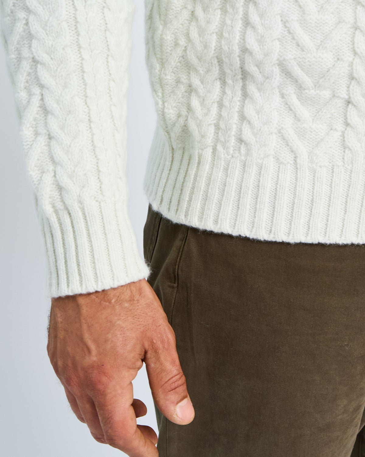 Close-up of a person wearing a white cable knit sweater and brown pants on a light gray background