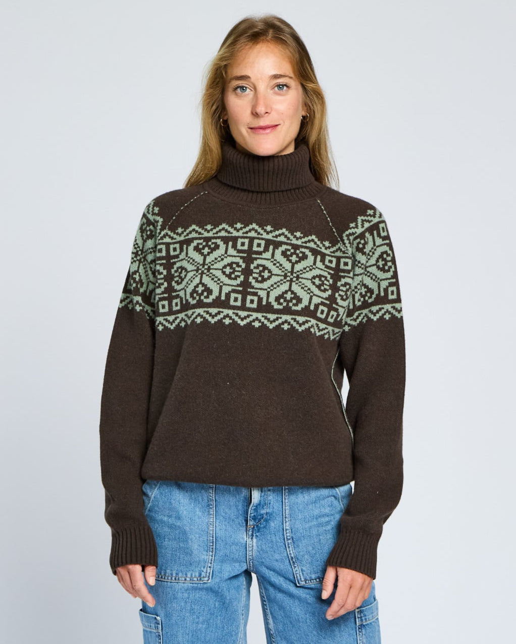 Woman wearing a brown sweater with green pattern and blue jeans on a light gray background