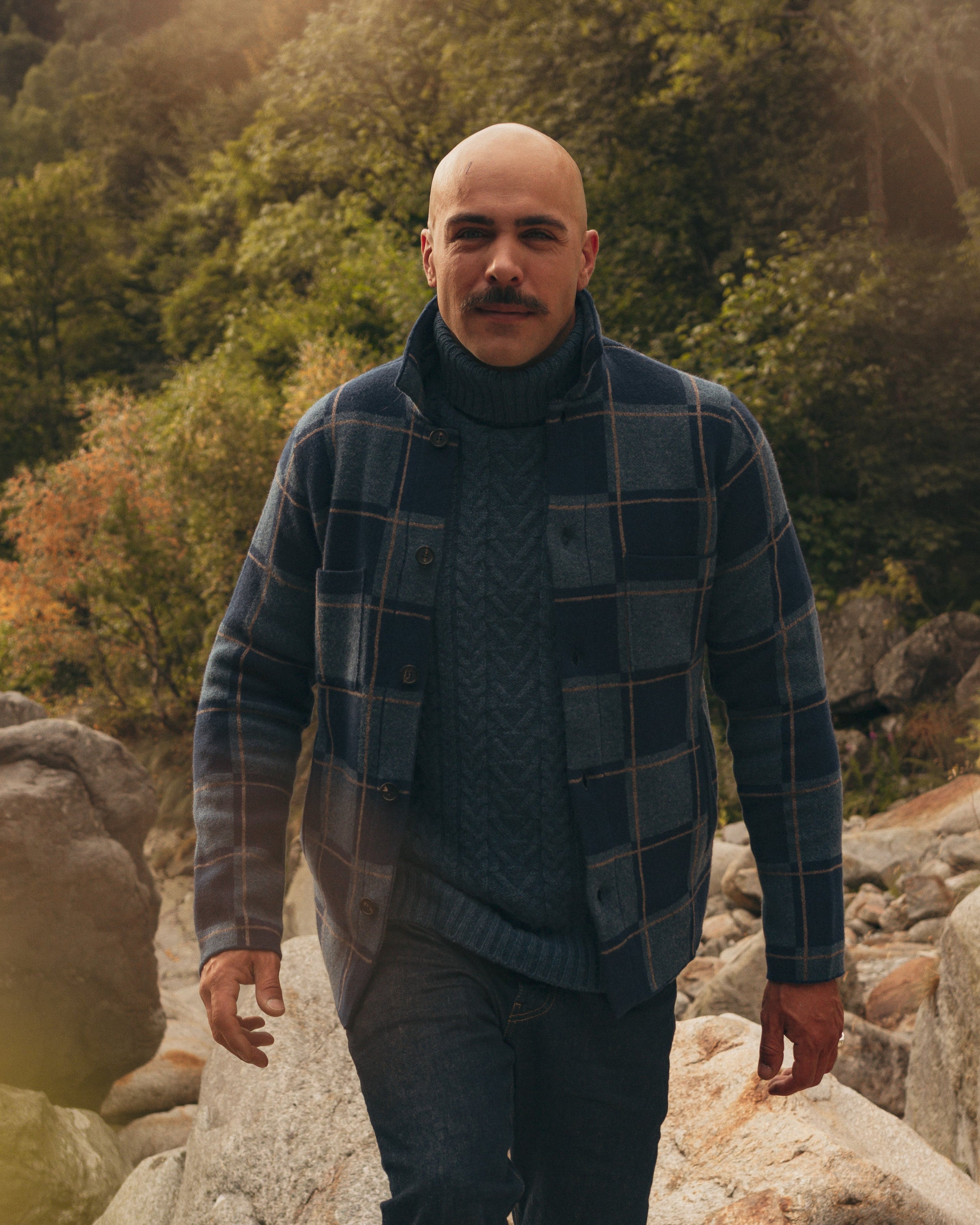 Man standing on rocks in a forested area wearing a plaid jacket and sweater.