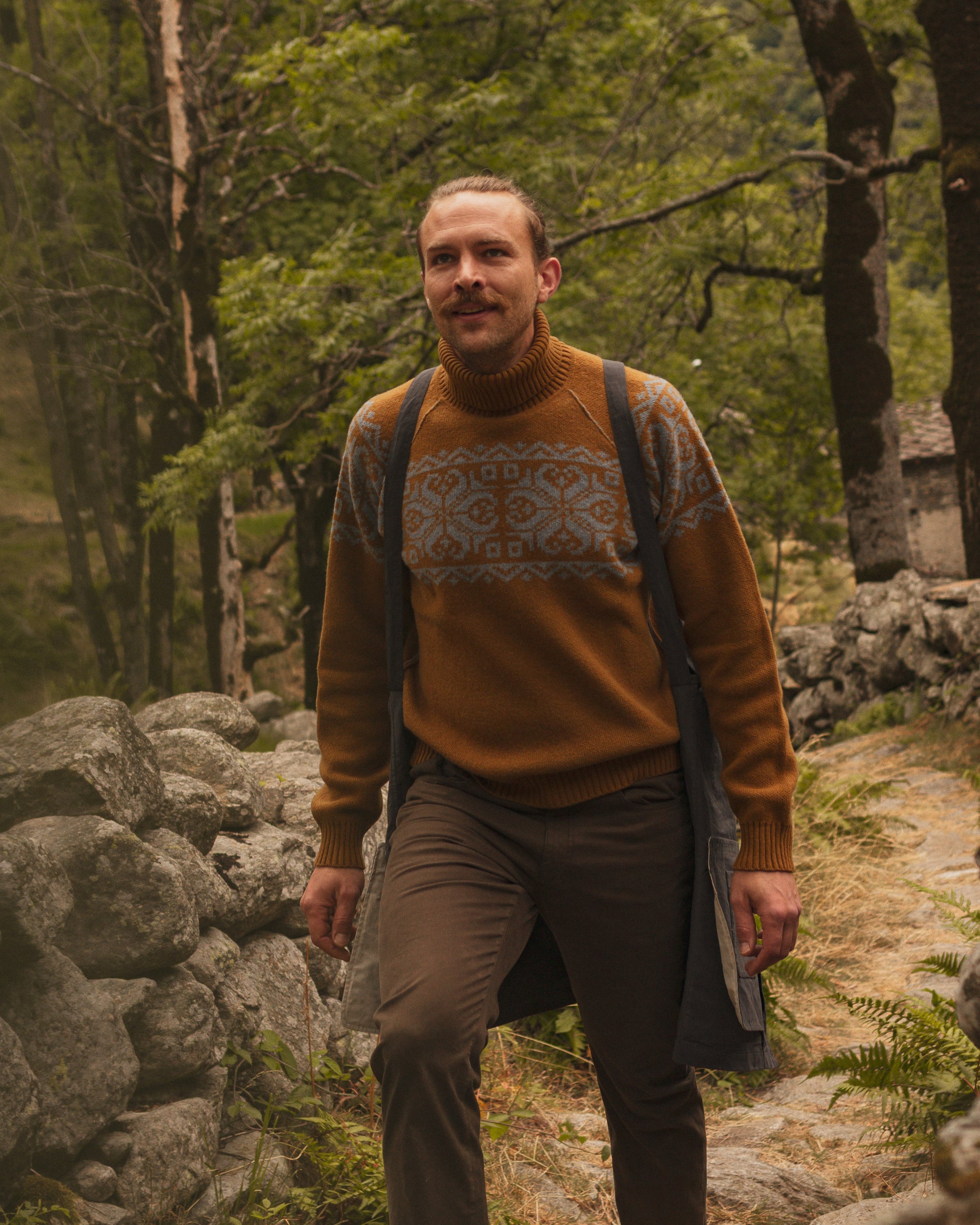 Man in a patterned sweater standing in a forest with stone walls.
