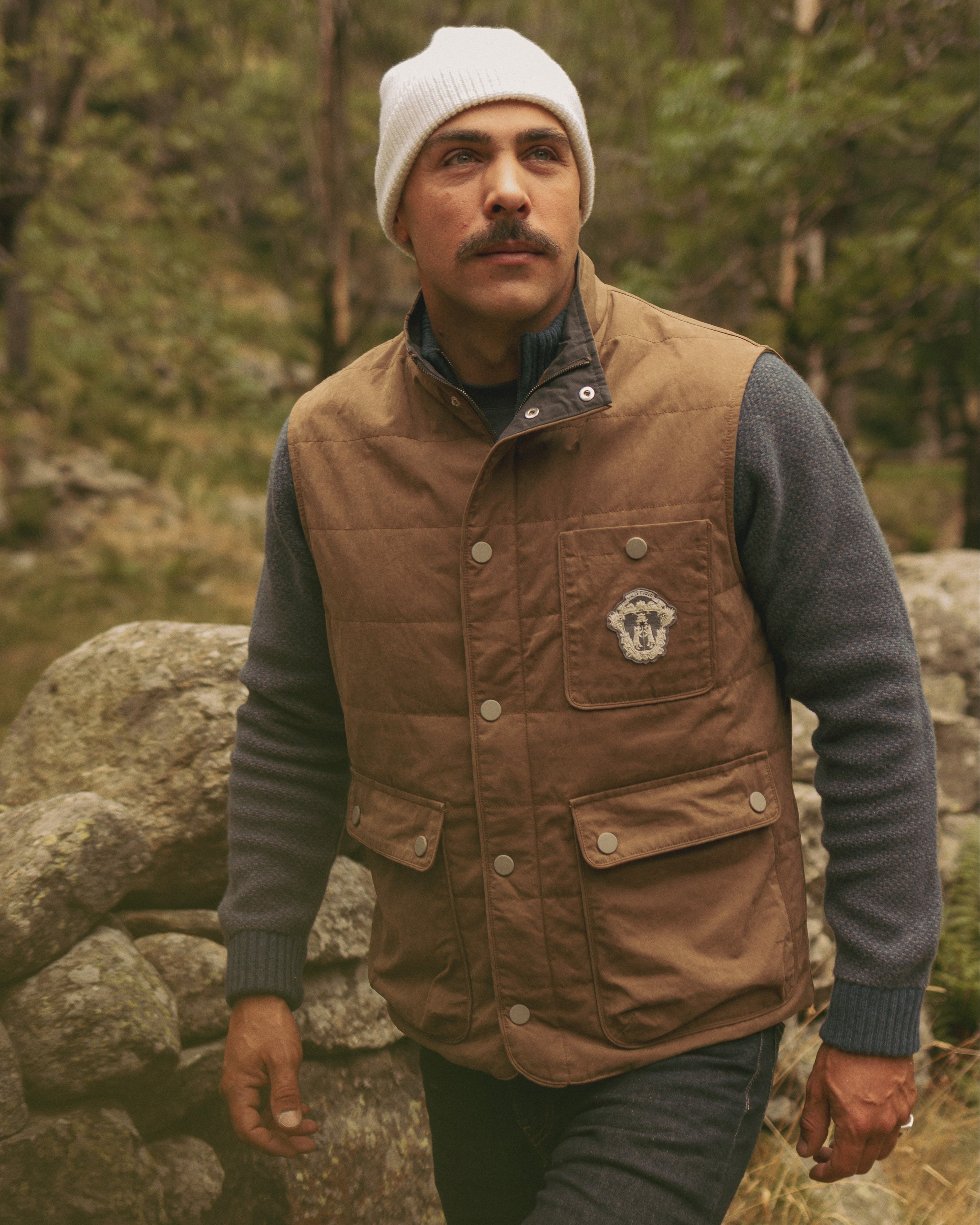 Man wearing a brown vest and white beanie standing in a forest.