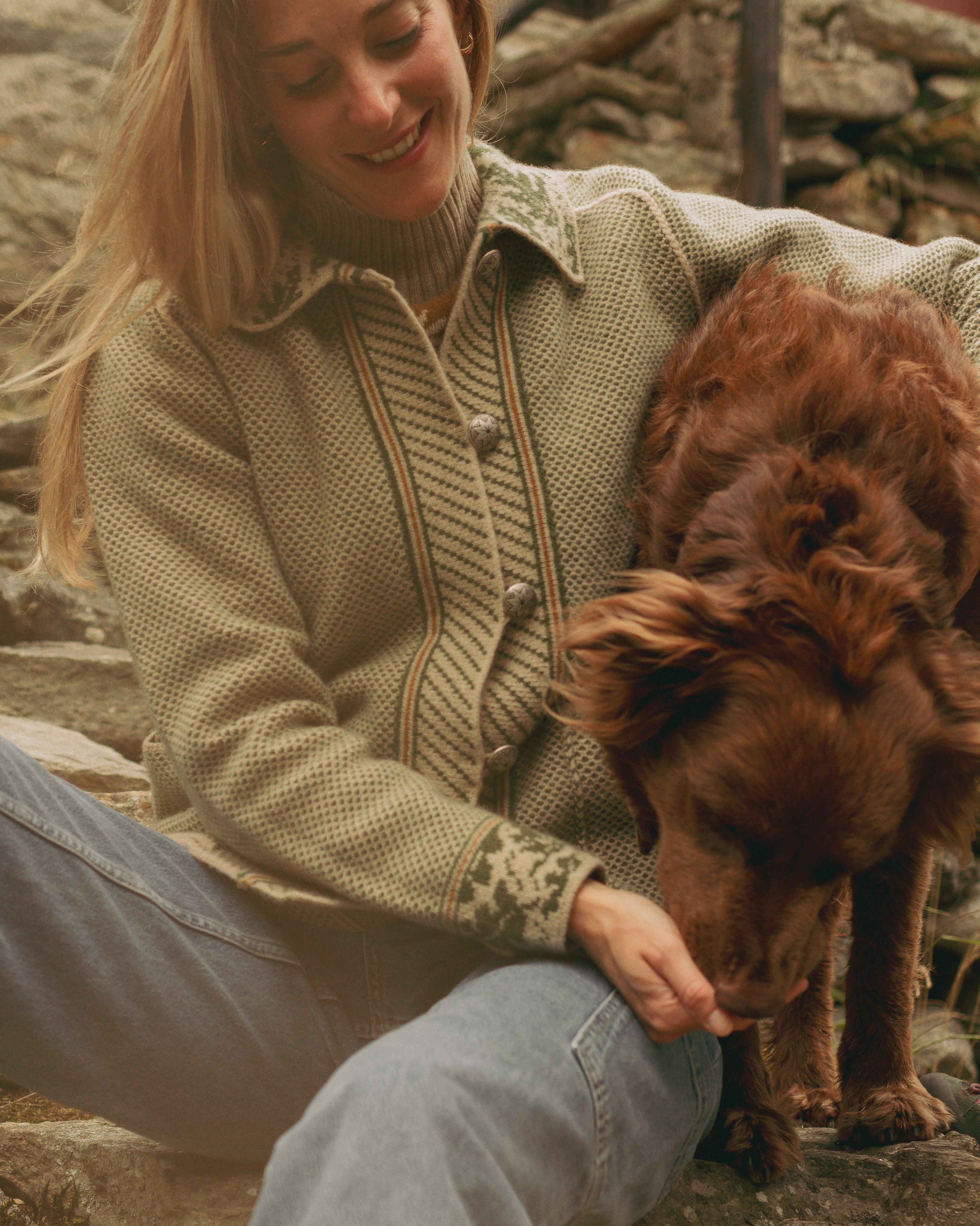 Woman in a green jacket sitting on rocks with a brown dog.
