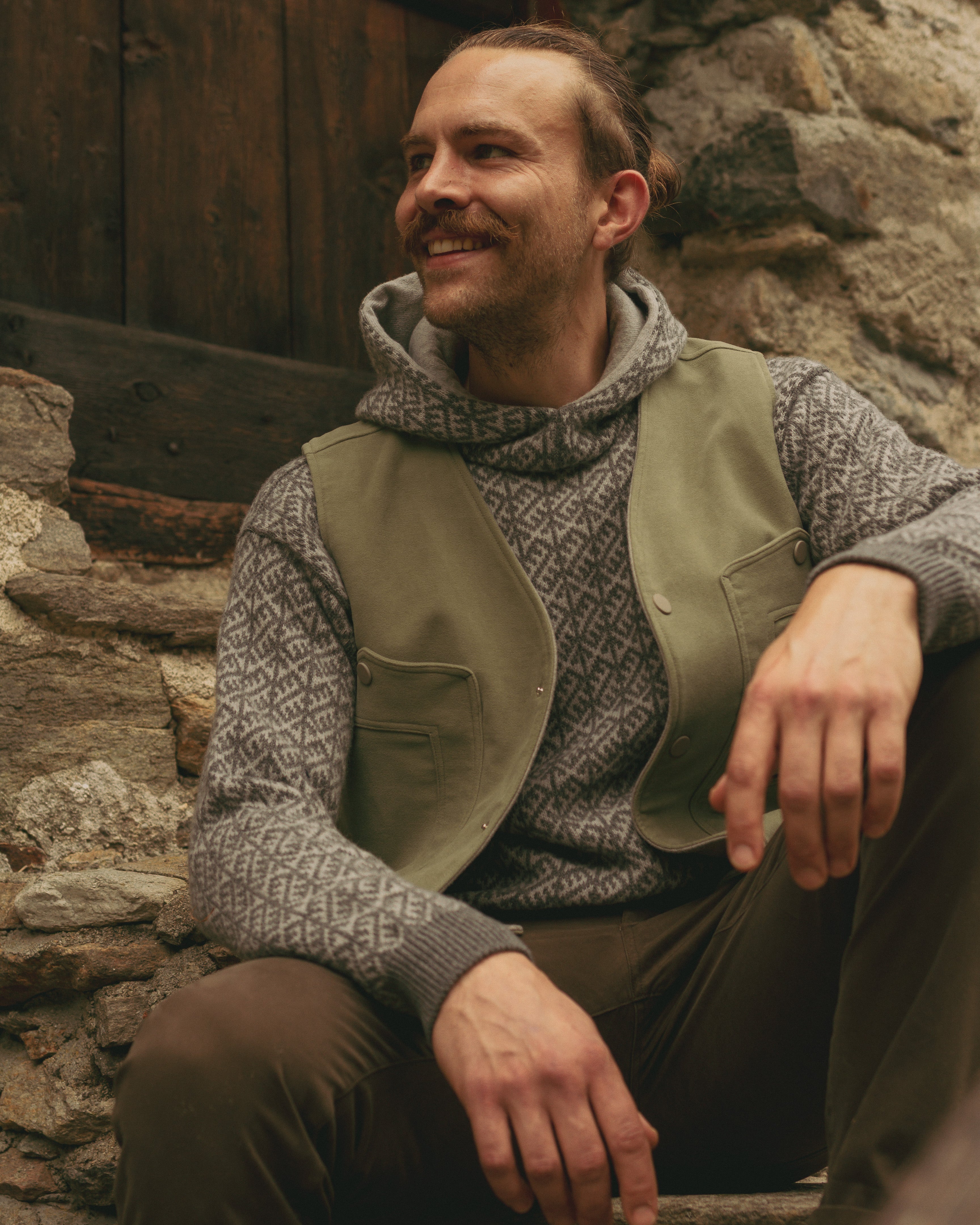 Man sitting on stone steps wearing a patterned sweater and green vest.
