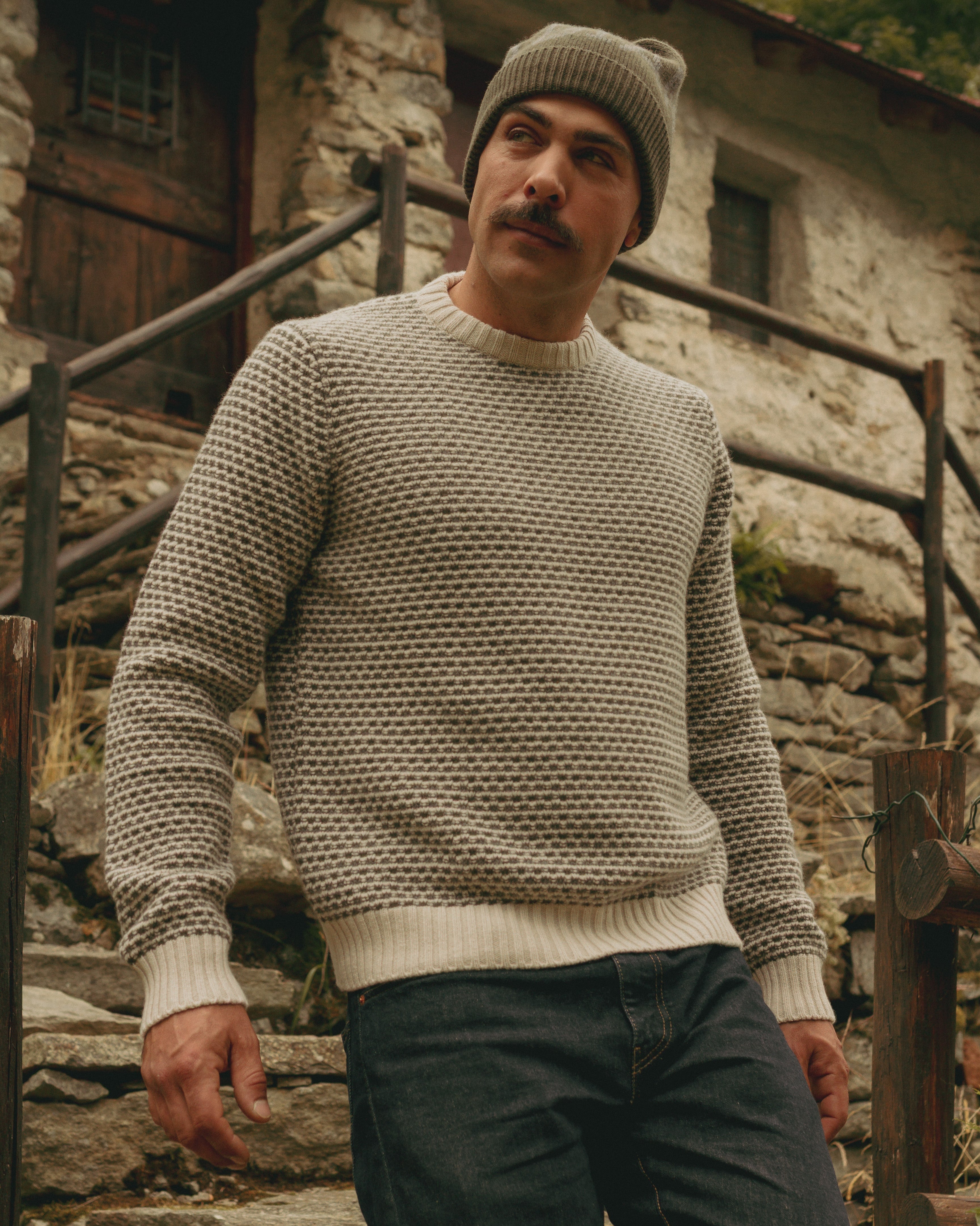 Man wearing a textured sweater and knit hat standing on stone steps with a rustic building in the background.