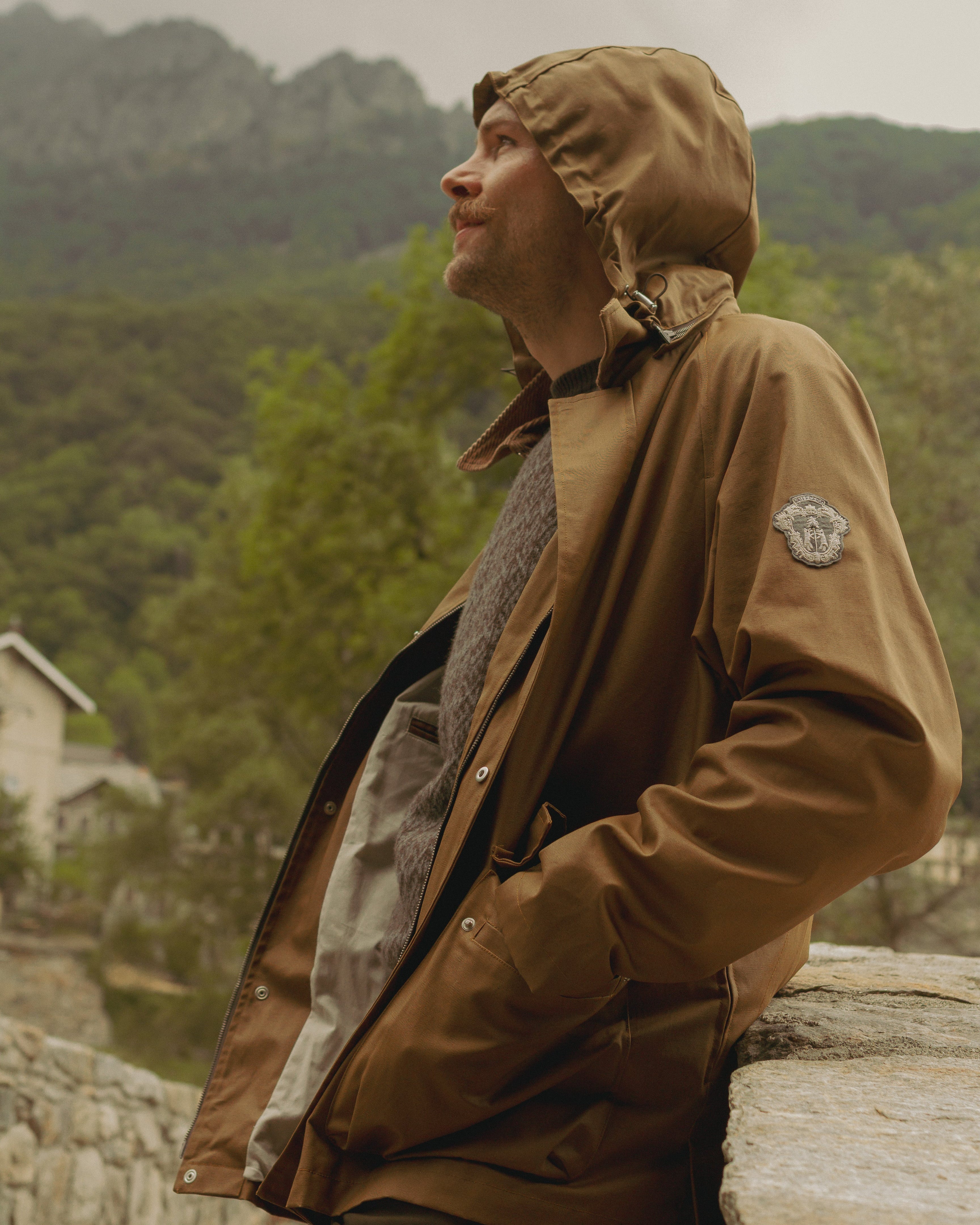 Man wearing a brown raincoat with a hood, standing outdoors with greenery in the background.