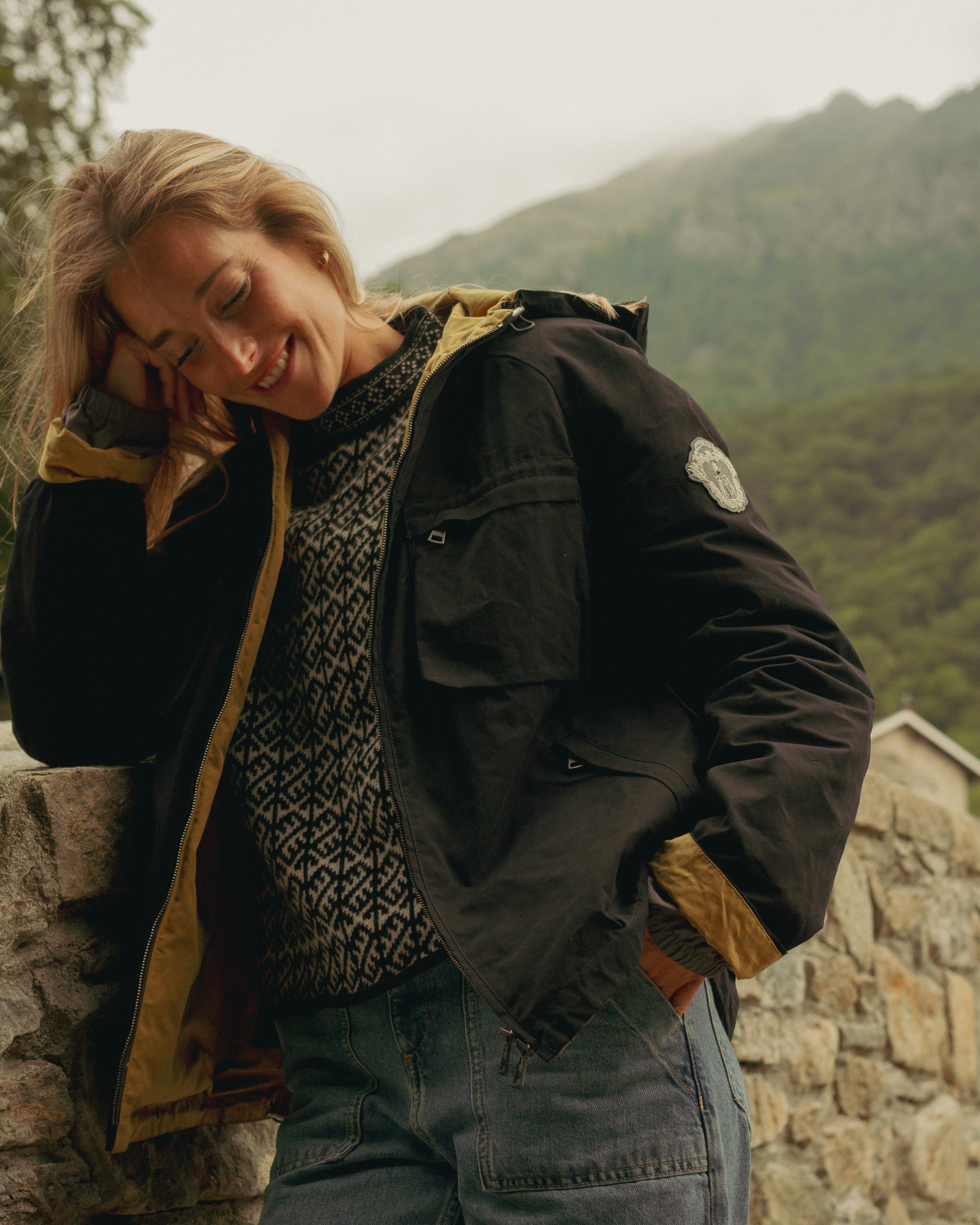 Person wearing a dark jacket with a yellow hood leaning against a stone wall with mountains in the background