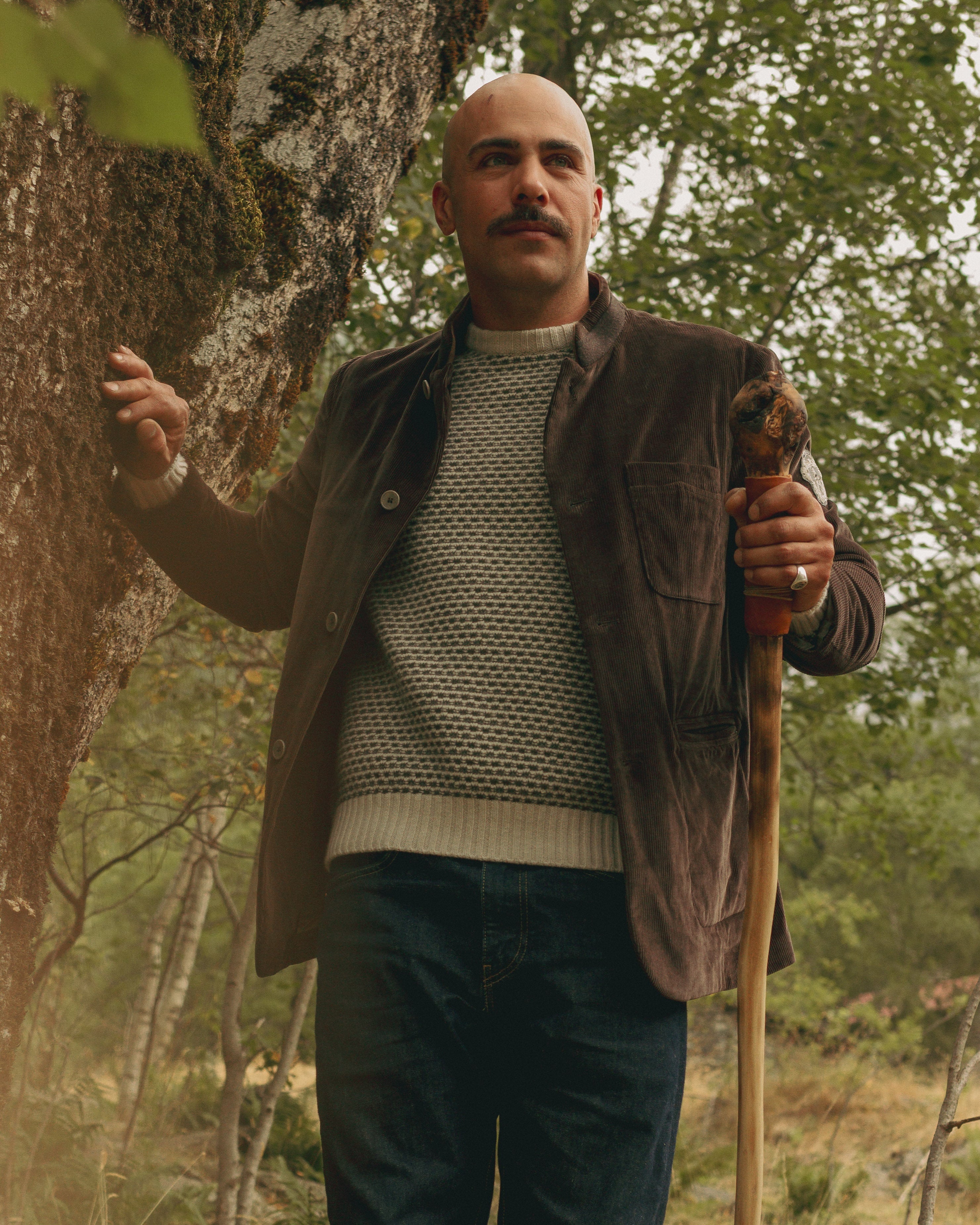 Man standing in a forest holding a walking stick, wearing a brown jacket and green sweater.